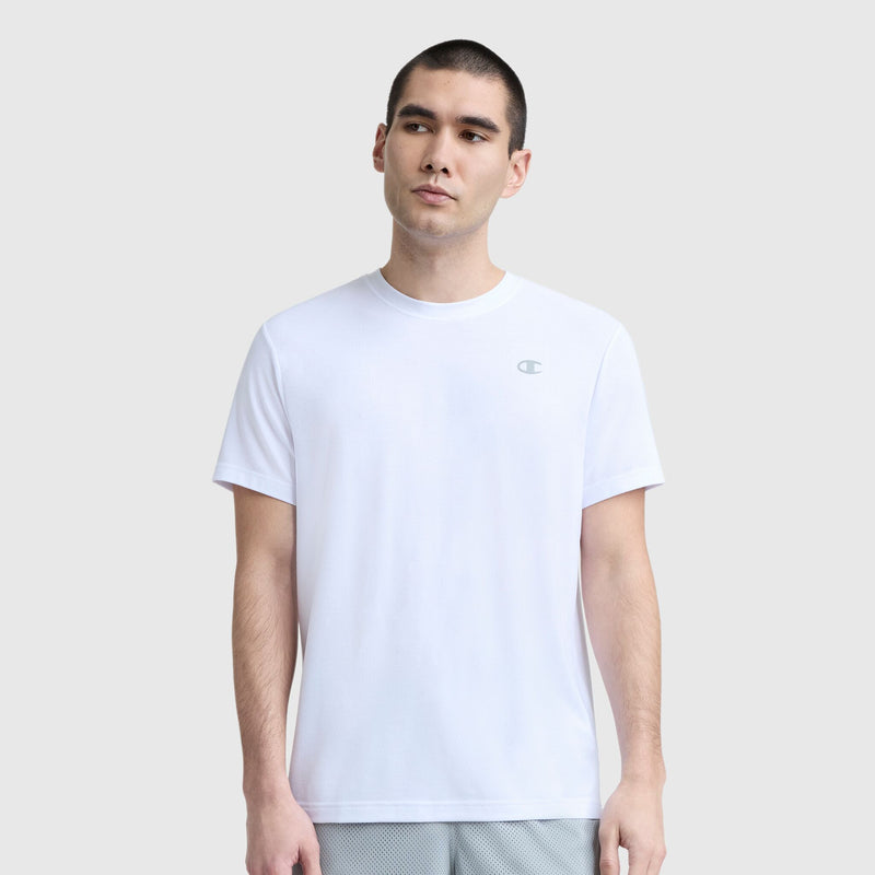 A man with short dark hair wears a Champion Sport Crewneck T-Shirt, Script Logo—a plain white men's slim-fit tee—paired with light gray shorts. He stands against a light gray background, looking slightly to the side.