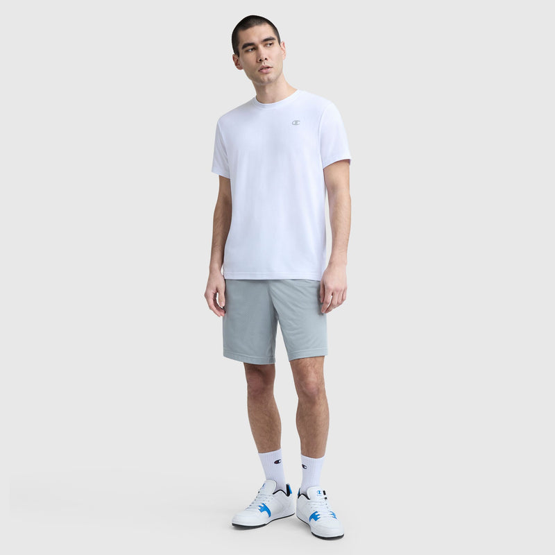 A man with short hair and a neutral expression stands against a light gray background, wearing a Champion Sport Crewneck T-Shirt with Script Logo (white), light gray shorts, white crew socks, and white sneakers with blue accents.