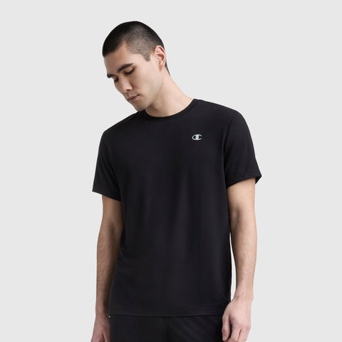 A young man with short dark hair wears a Champion Sport Crewneck T-Shirt, Script Logo, standing against a light gray background and looking down with a neutral expression.