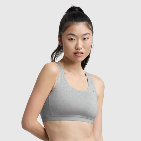 A woman with long dark hair in a ponytail wears a Champion Compression Sports Bra, C Logo, in light gray and looks at the camera against a plain light gray background.