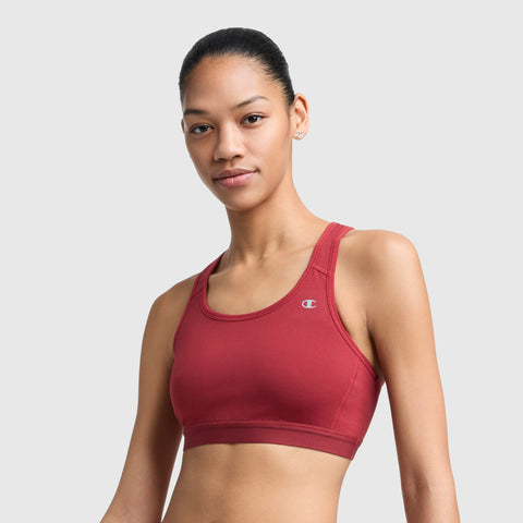 A woman with pulled-back hair wears a red Champion Compression Sports Bra, C Logo, offering moderate support. She stands against a plain light gray background, looking at the camera with a slight smile.