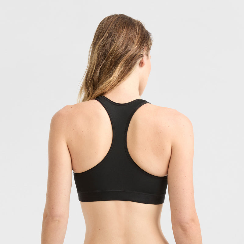 A woman with long, light brown hair is seen from the back wearing the Champion Compression Sports Bra, C Logo, in black against a plain light gray background.