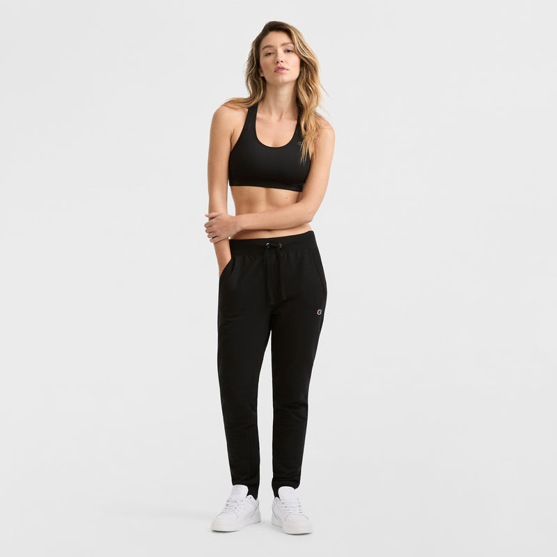 A woman stands against a white background wearing the Champion Compression Sports Bra, C Logo, paired with black jogger pants and white sneakers, her right arm resting across her body and a neutral expression on her face.