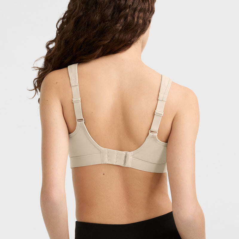 A woman with long, wavy brown hair is shown from the back wearing a beige Champion Spot Comfort High-Impact Sports Bra, C Logo, with wide straps for maximum support and black bottoms against a plain light background.