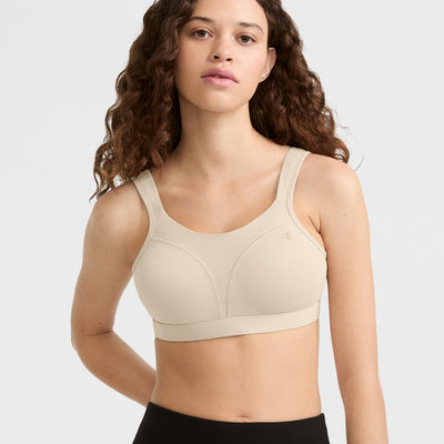 A woman with long, curly brown hair wears the Champion Spot Comfort High-Impact Sports Bra, C Logo in beige and black leggings, standing against a plain light gray background.