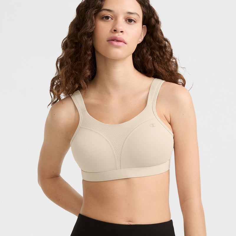 A woman with long, curly brown hair wears the Champion Spot Comfort High-Impact Sports Bra, C Logo in beige and black leggings, standing against a plain light gray background.