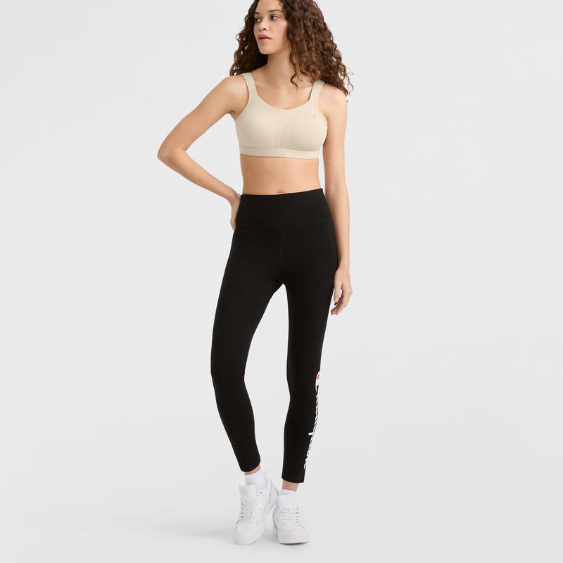 A woman with long curly hair models the Champion Spot Comfort High-Impact Sports Bra (C Logo) in beige, paired with high-waisted black leggings featuring white text on the left leg and white sneakers, standing against a plain white background.