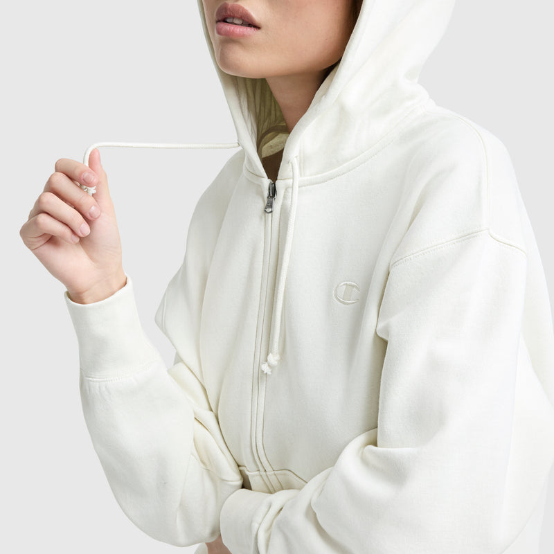 A person in a Champion Powerblend Fleece Zip-Up Hoodie, C Logo, off-white cotton blend, wears the hood up and holds one drawstring; only the lower part of their face is visible.