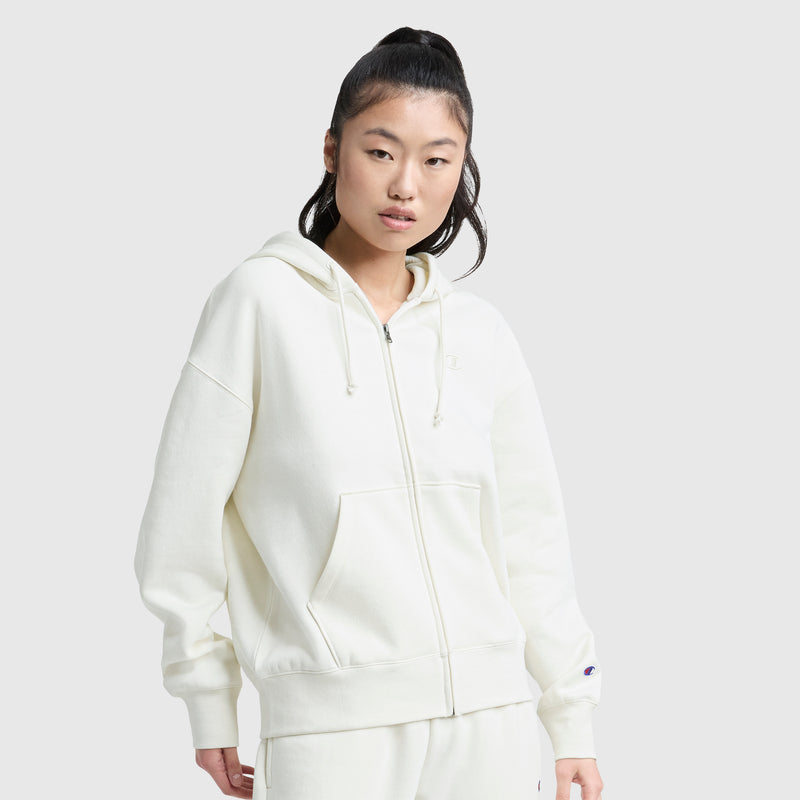 A person with long dark hair in a high ponytail wears a Champion Powerblend Fleece Zip-Up Hoodie, C Logo, paired with matching pants, standing against a plain light gray background.