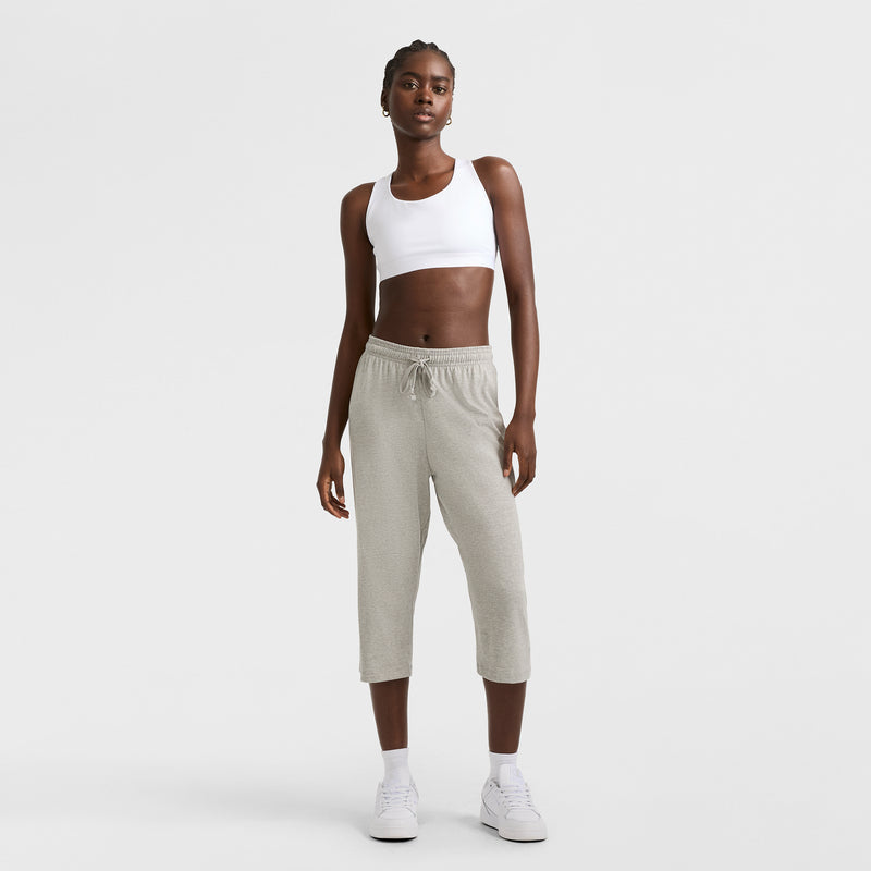 Against a white background, a woman models Champion's Lightweight Lounge Capri Pants, C Logo, in light gray with a white sports bra and sneakers. She stands relaxed and confident, looking directly at the camera.