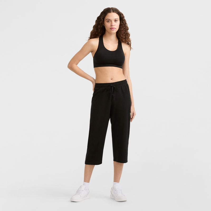 A woman with long curly hair models Champion Lightweight Lounge Capri Pants (C Logo) in black, paired with a black sports bra, white socks, and white sneakers against a plain light gray background.