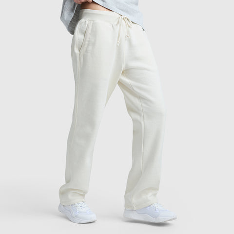 A person wears cream-colored Champion Powerblend Straight Hem Sweatpants made from midweight fleece and white sneakers, standing against a plain light background. The shot focuses on the lower body with hands in pockets and a straight hem finish.
