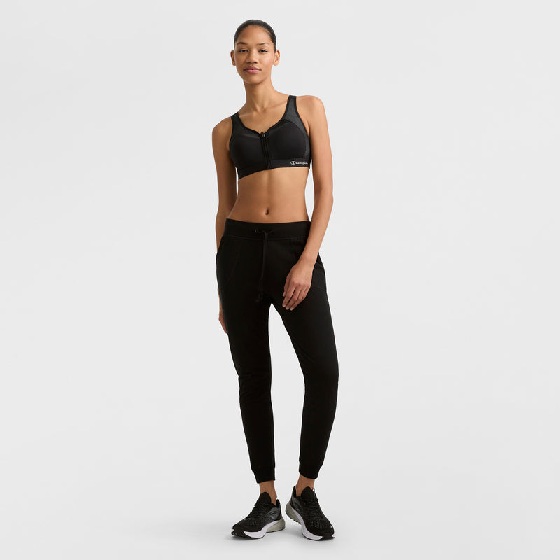 A woman stands against a white background, wearing Champion Lightweight Lounge Joggers, a black sports bra, and black sneakers. She poses casually with one hand at her side and a subtle smile.