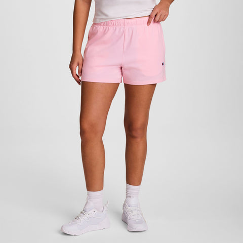 A person wears light pink Champion Practice Shorts, a white t-shirt, white socks, and white sneakers while standing against a plain, light background.