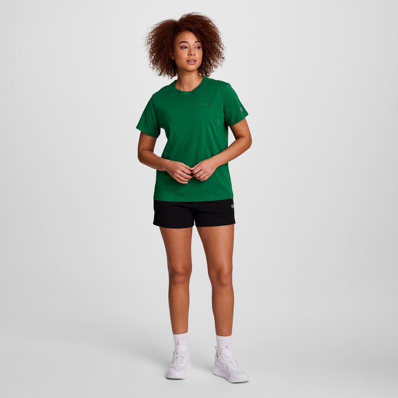 A person with curly hair stands against a white background, facing forward with hands loosely clasped, wearing a green t-shirt and Champion Practice Shorts for women, paired with white socks and white sneakers.