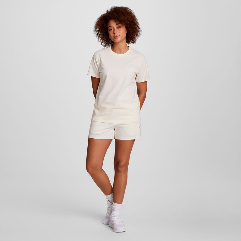 A woman with curly hair stands against a plain light background, wearing a cream t-shirt, Champion Practice Shorts, white socks, and white sneakers. She has her hands behind her back and appears relaxed.