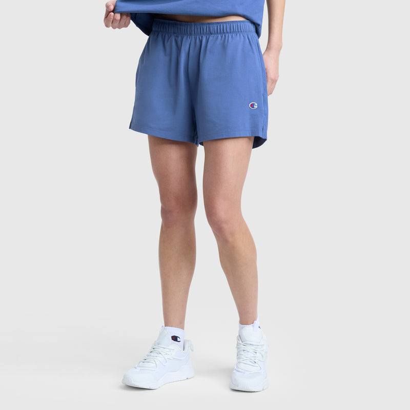 A person is shown from the waist down, wearing Champion Practice Shorts in light blue with an elastic waistband, paired with a matching blue shirt and white sneakers, standing against a plain light gray background.