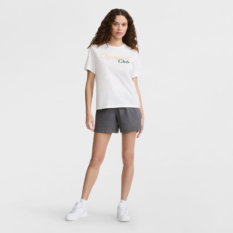 A woman with curly brown hair wears a white T-shirt with Champions Club text, Champion 5" Lightweight Lounge Shorts with a C logo in dark gray, and white sneakers, standing against a plain white background.