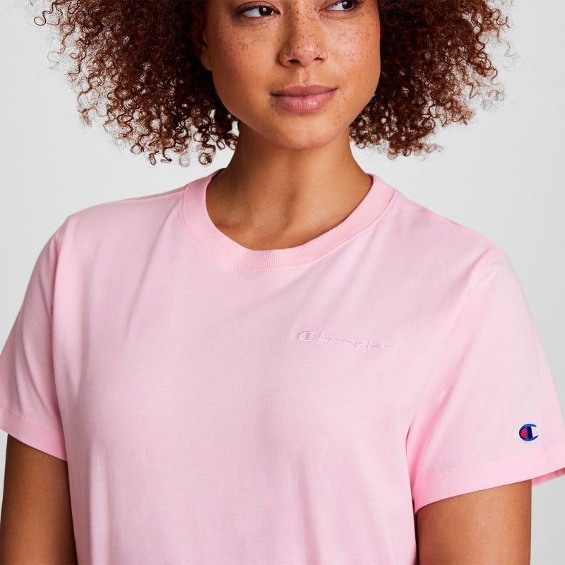 A person with curly brown hair wears a light pink Champion Classic T-Shirt, featuring tonal left chest script embroidery and a small Champion patch on the left sleeve against a plain, neutral background.