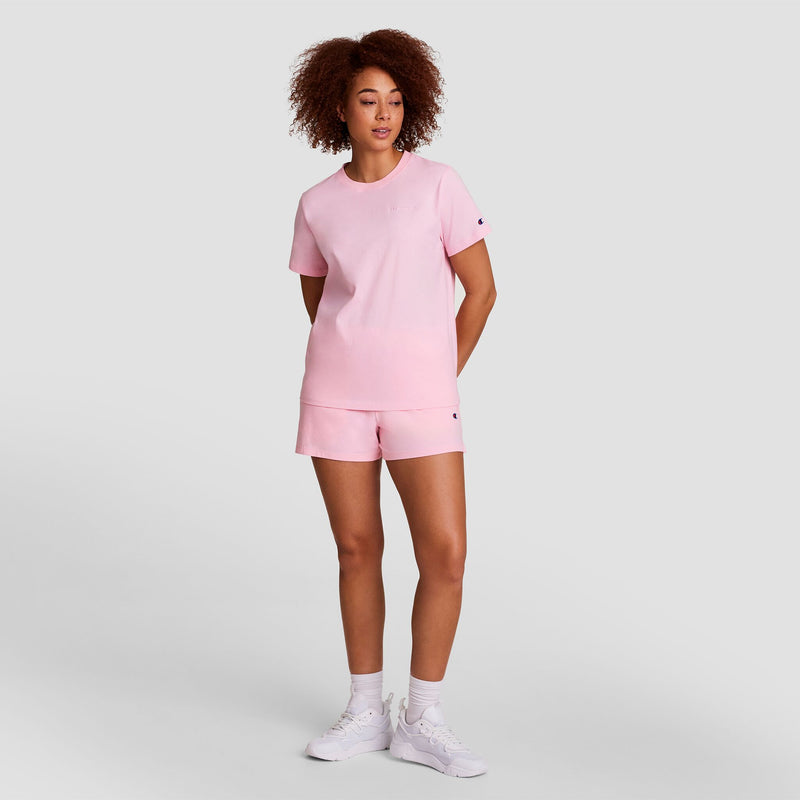 A woman with curly hair wears the Champion Classic T-Shirt, Tonal Left Chest Script in light pink, paired with matching shorts, white socks, and sneakers. She stands against a plain gray background with her hands behind her back.