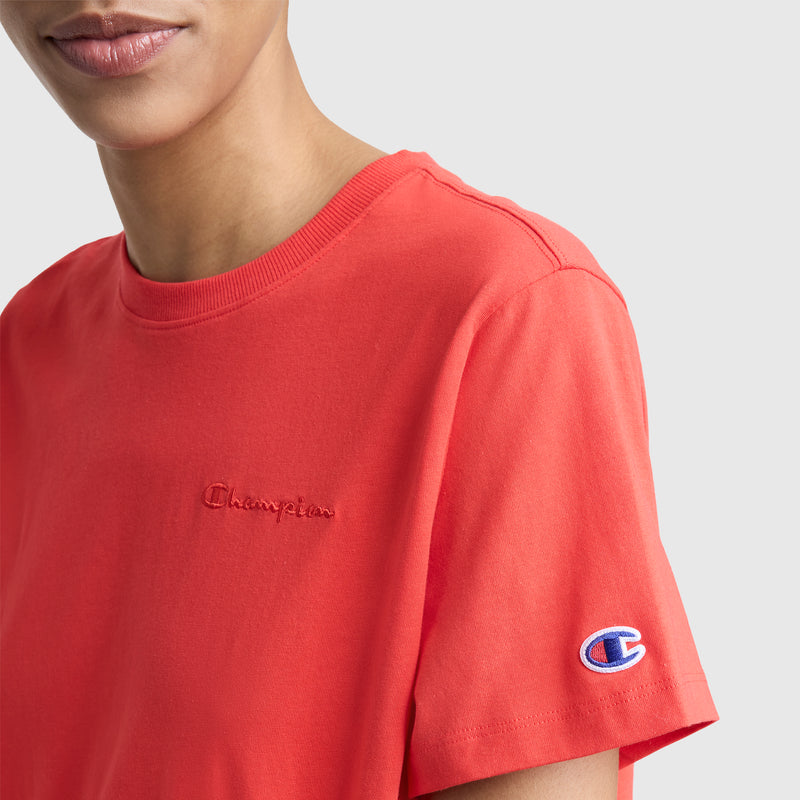 A person shown from the shoulders up wears a bright red Champion Classic T-Shirt featuring tonal script on the left chest, crafted from a soft cotton blend. An embroidered C logo patch accents the left sleeve.