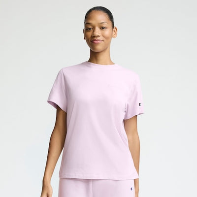 A person stands facing forward, smiling slightly, wearing a light pink short-sleeve t-shirt and matching pants against a plain light background.