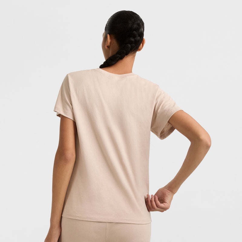 A person with a long braid is shown from the back, wearing a beige Champion Classic T-Shirt with tonal left chest script and matching pants, standing against a plain light background.