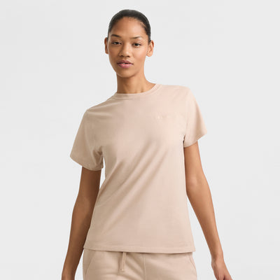 A woman wears the Champion Classic T-Shirt with Tonal Left Chest Script in beige, paired with matching beige pants, standing against a plain light background and looking at the camera with a relaxed expression.