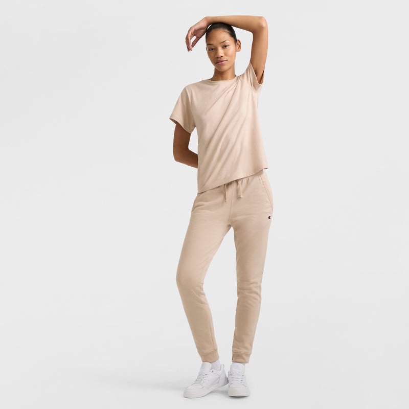 A person stands against a plain white background, wearing the Champion Classic T-Shirt with Tonal Left Chest Script, paired with beige joggers and white sneakers. One arm is bent above their head for a relaxed, casual vibe.