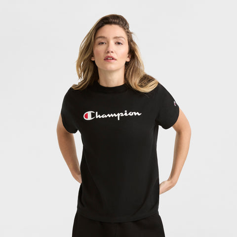 A woman with long, light brown hair wears the Champion Classic T-Shirt, Script Logo, standing against a plain light gray background with her hands behind her back.