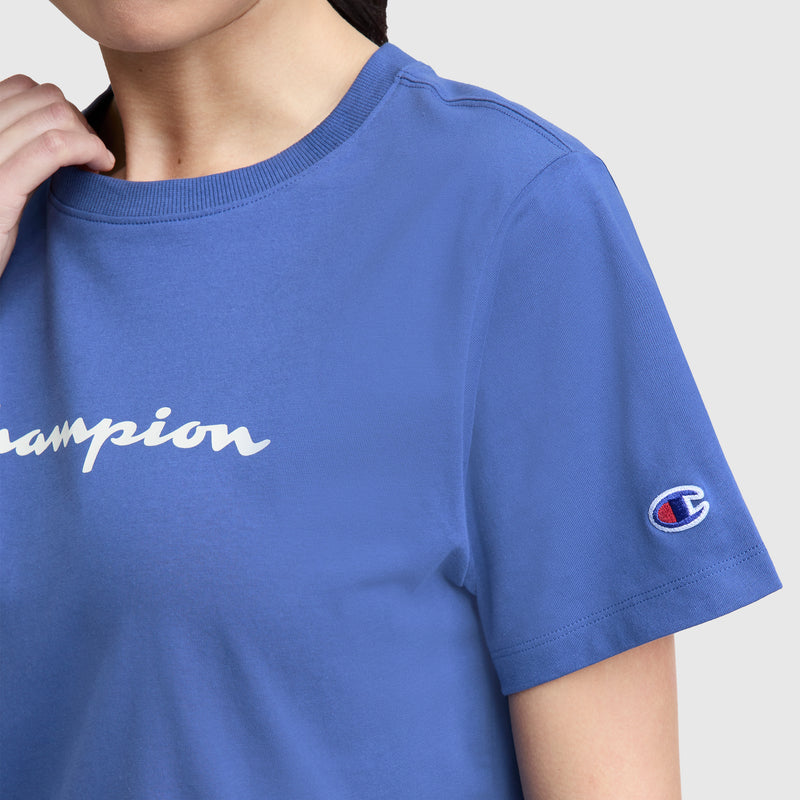 A person wears a blue Champion Classic T-Shirt, Script Logo, with retro script partially visible on the front and a small sleeve logo patch. Only the lower face, neck, and upper torso are shown.