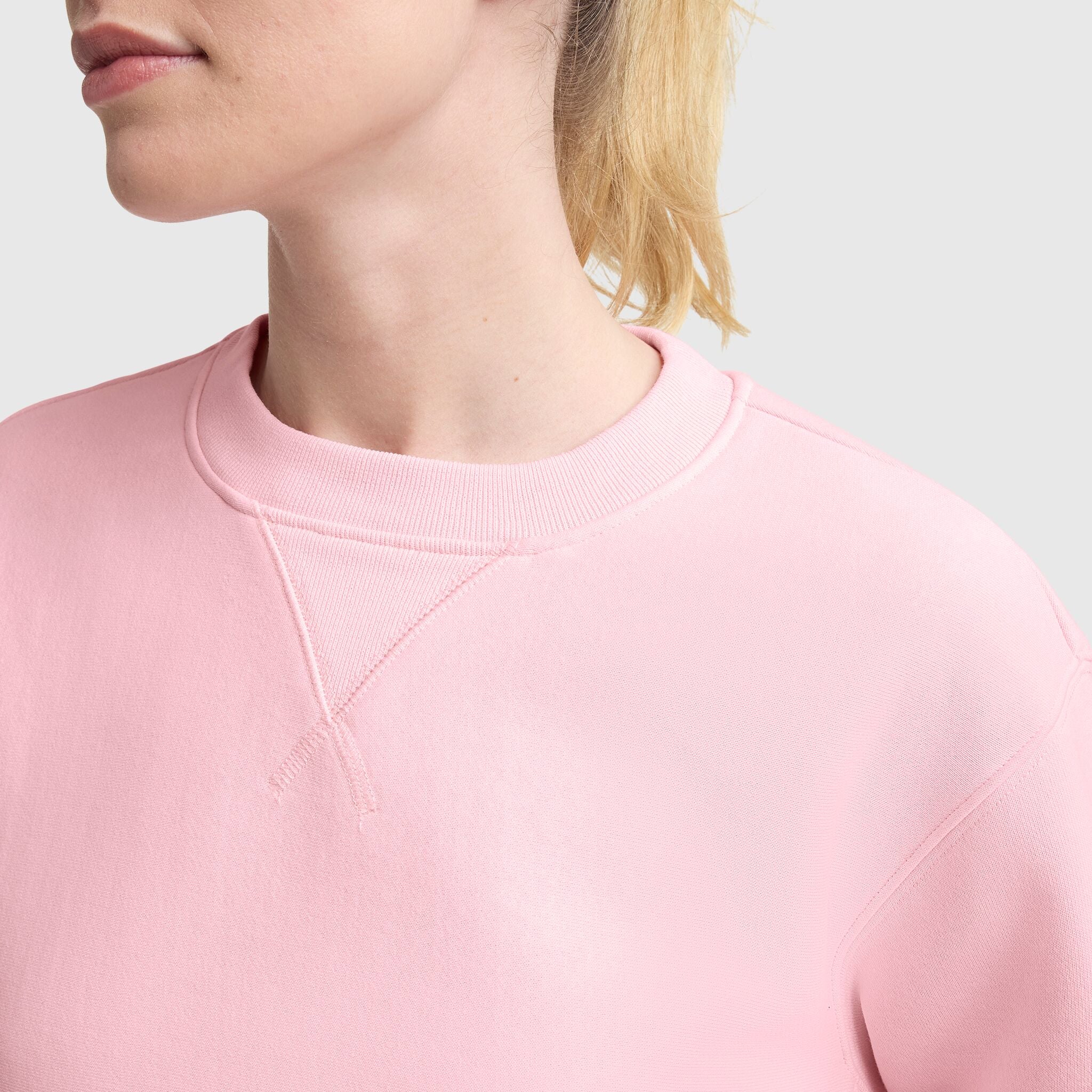 A light-skinned, blonde-haired person wears a Champion Essentials Reverse Weave Crew Neck Sweatshirt in light pink. The image is cropped to show the lower face, neck, and upper torso against a plain light gray background.