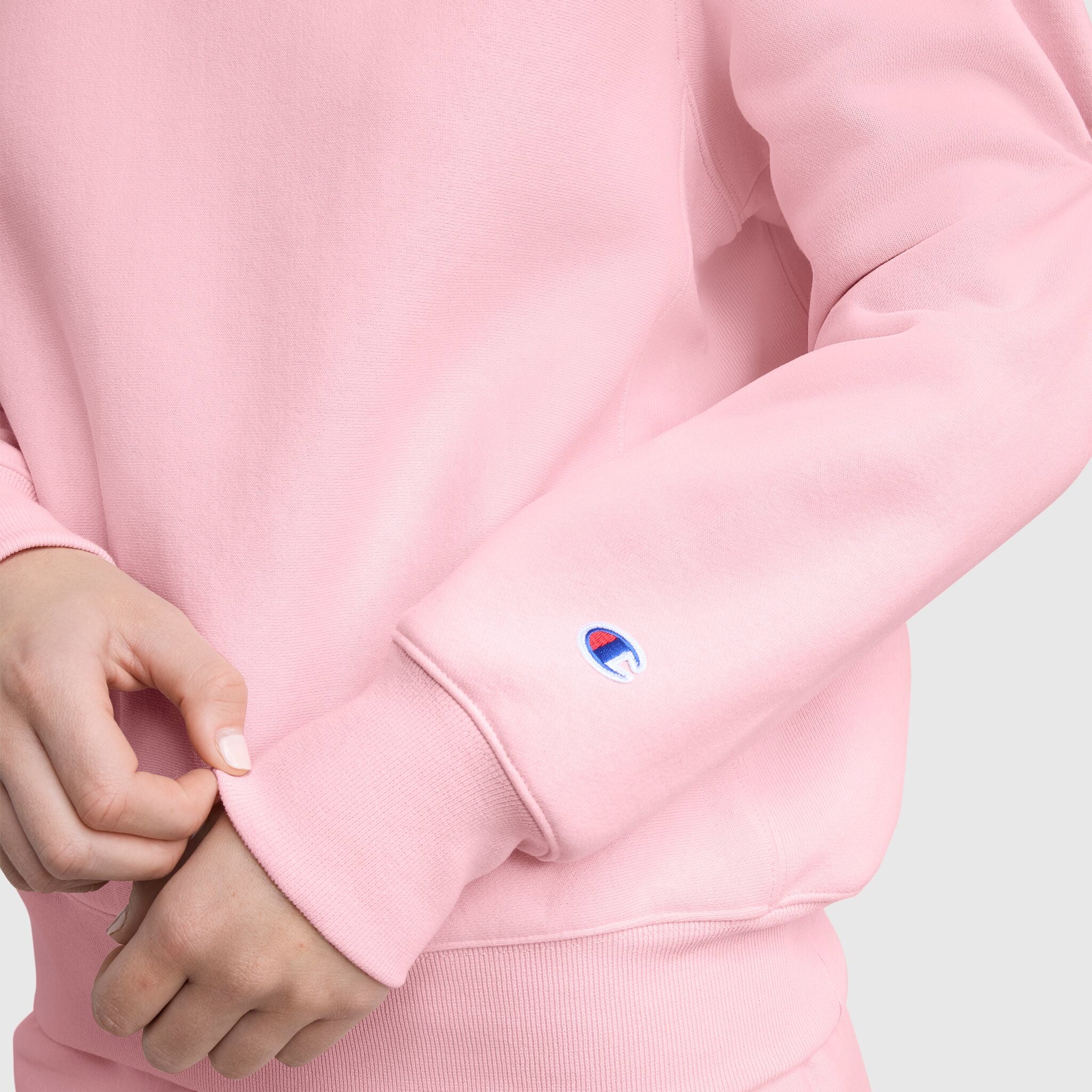 A person wearing a light pink Champion Essentials Reverse Weave Crew Neck Sweatshirt adjusts the sleeve, revealing a small embroidered blue, red, and white C logo near the wrist. The heavyweight fleece is shown against a plain, light-colored background.