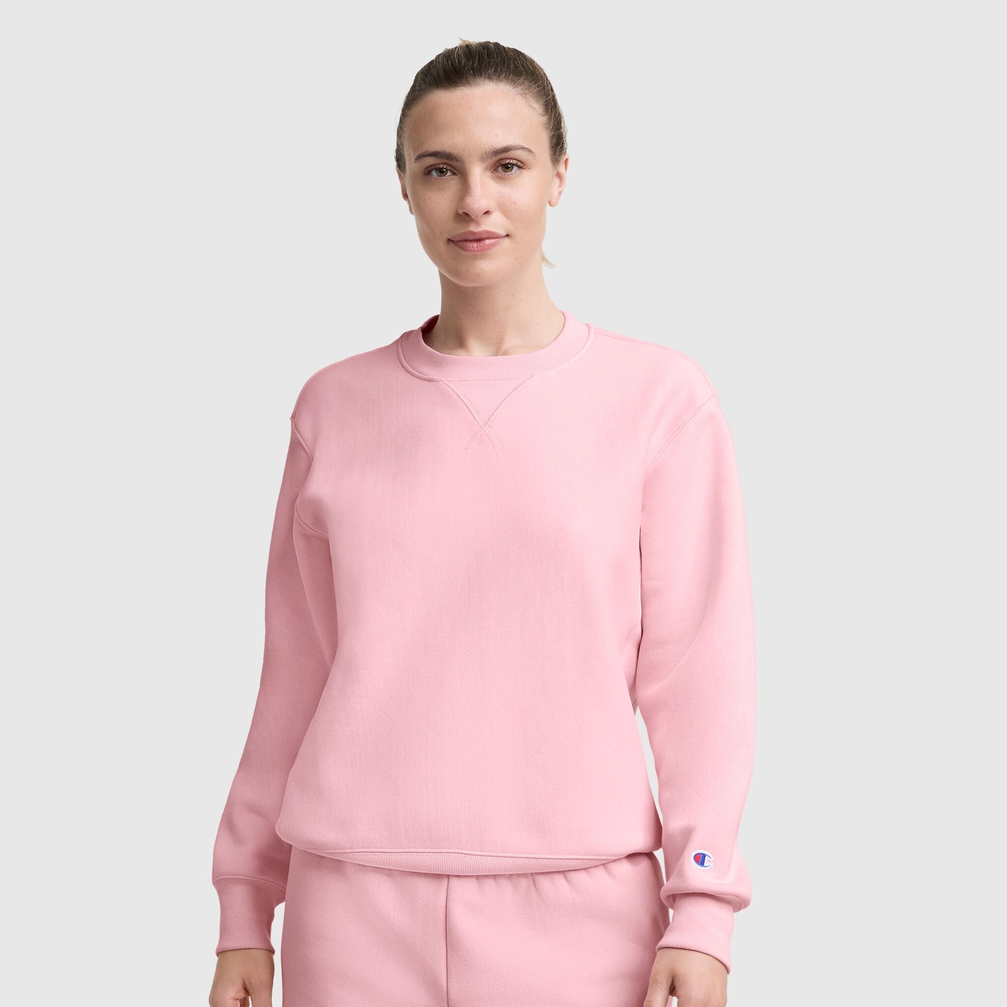 A woman with light brown hair tied back wears a Champion Essentials Reverse Weave Crew Neck Sweatshirt and matching pants, standing against a plain light gray background. She faces forward with a neutral expression, reflecting classic comfort.