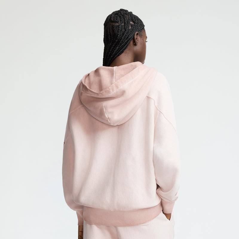 A person with braided hair stands with their back to the camera, wearing a light pink Champion Time Capsule Vintage 80's Full Zip Hoodie and matching pants against a plain, light background.