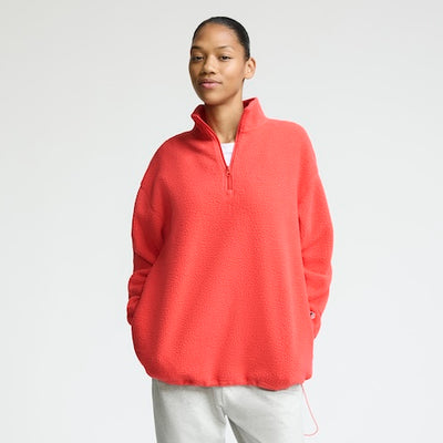 A person stands facing forward, wearing a bright coral red fleece pullover with a front zipper and light gray pants, against a plain light background. They have short hair and a relaxed, confident expression.
