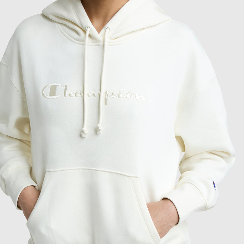A person wearing an off-white Champion Tonal Script Freshman Powerblend Hoodie with hands in the front pocket. The women's pullover features the Champion logo embroidered across the chest.
