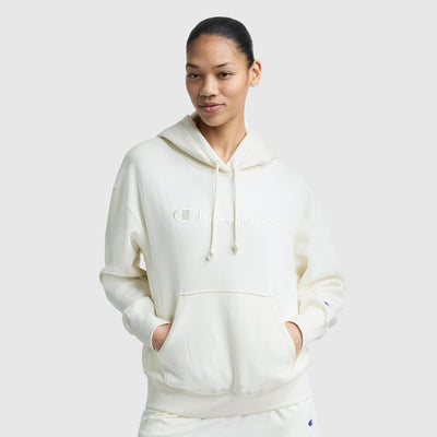 A person wearing a Champion Tonal Script Freshman Powerblend Hoodie in off-white stands against a plain light gray background, hands in the front pocket, looking forward with a slight smile.