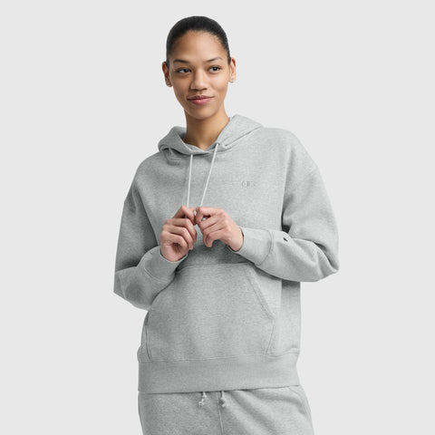 Wearing a light gray Champion Small C Freshman Powerblend Hoodie with an adjustable drawcord hood and matching sweatpants, a person stands against a plain backdrop, looking off to the side with a slight smile and hands holding the hoodie strings.