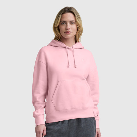 A person with shoulder-length blonde hair wears the Champion Small C Freshman Powerblend Hoodie in light pink, featuring midweight fleece, front pocket, and adjustable drawcord hood, paired with gray pants against a plain light gray background.