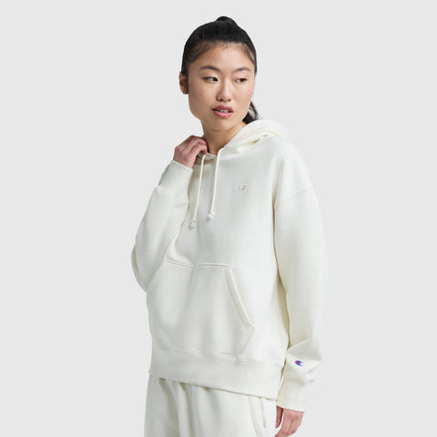 A person with long dark hair in a ponytail wears the Champion Small C Freshman Powerblend Hoodie, off-white and loose-fitting, with matching sweatpants, standing against a plain light gray background and looking slightly to the side.