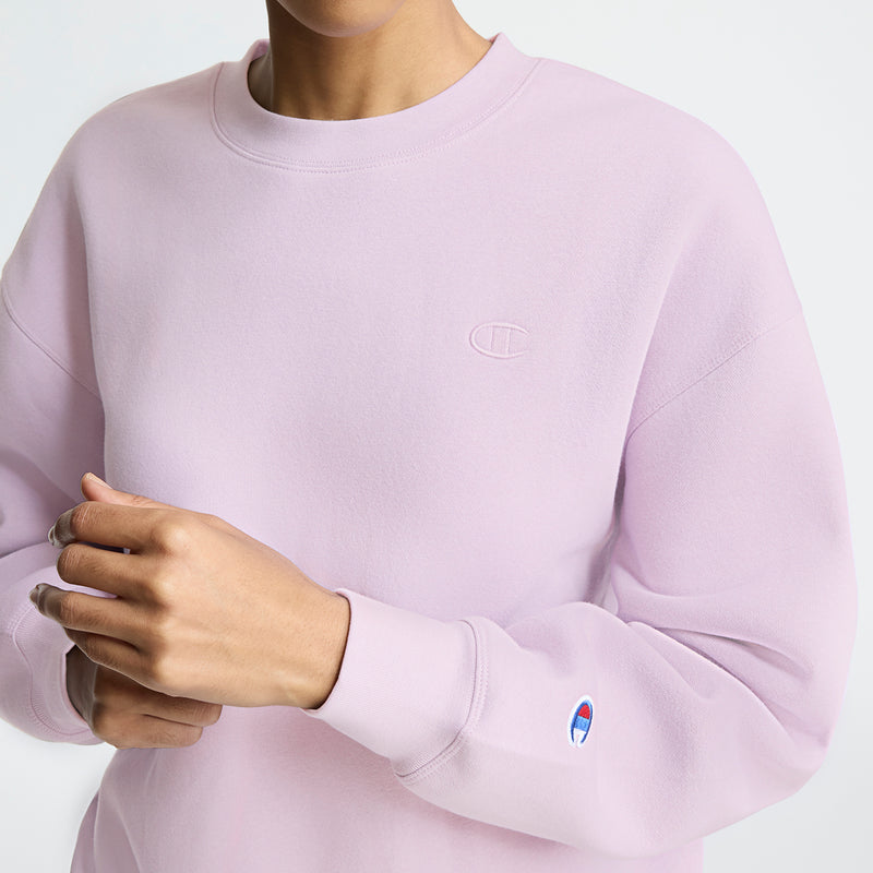 A person wearing a light pink Champion Powerblend Crewneck sweatshirt, featuring the small embroidered "C" logo on the chest and sleeve. Made with midweight fleece for comfort. The person's hands, with neutral nail polish, are visible.