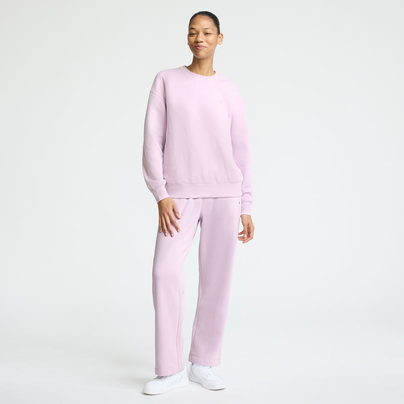 A person stands against a white background, wearing the Champion Powerblend Crewneck with a small "C" logo and matching sweatpants, paired with white sneakers. They smile warmly, embodying everyday comfort in midweight fleece.