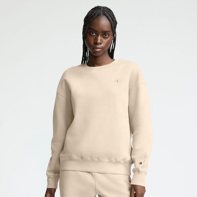 A woman with long braids wears a beige crewneck sweatshirt and matching sweatpants, standing against a plain light background.