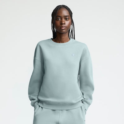 A woman with braided hair wears a light blue sweatshirt and matching sweatpants, standing against a plain light background with her hands in her pockets, looking directly at the camera.