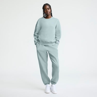 A person with long braids stands against a plain background, wearing a light blue sweatshirt and matching sweatpants with white sneakers. Their hands are in their pockets and they are looking at the camera.