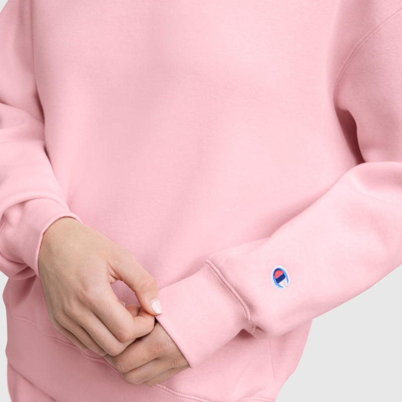 A person wears a light pink Champion Powerblend Crewneck Sweatshirt with an embroidered script logo, sleeves featuring the small Champion patch near the wrist. Their hands are folded at the waist against a plain, light-colored background.