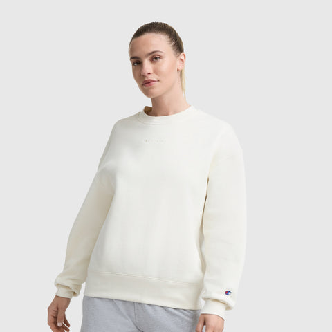 A person stands against a plain light gray background, wearing a loose-fit Champion Powerblend Crewneck Sweatshirt with an embroidered script logo and light gray pants, hair pulled back in a ponytail.