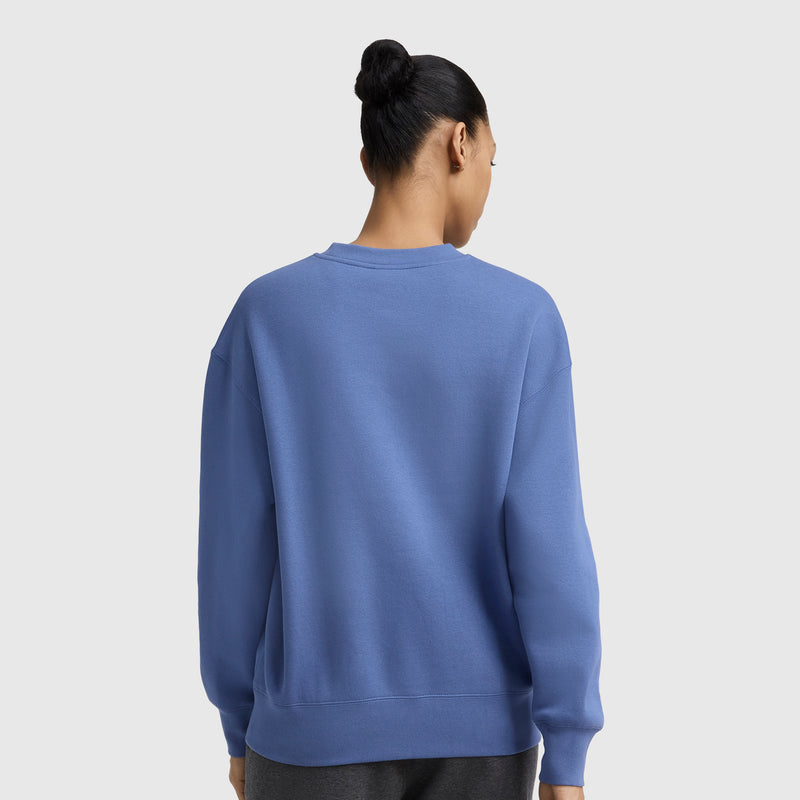 A person with dark hair in a bun is seen from behind wearing the Champion Powerblend Crewneck Sweatshirt with embroidered script logo and dark pants, standing against a plain light gray background.