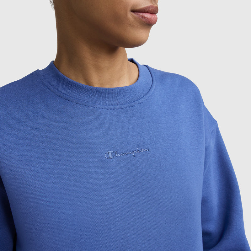 A woman wears the Champion Powerblend Crewneck Sweatshirt with an embroidered script logo, shown from the shoulders up against a plain background.